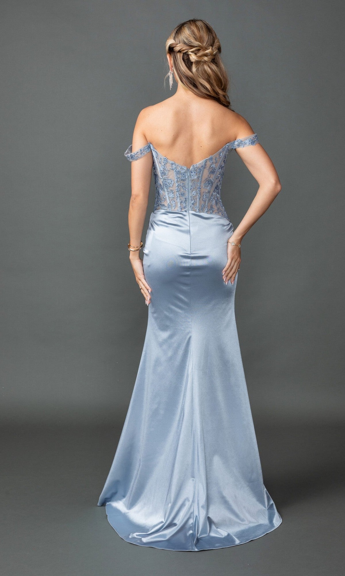 A woman faces away in the Long Prom Dress: Dancing Queen 4536, a light blue lace corset style with off-the-shoulder straps, a fitted satin skirt, and delicate lace bodice details against a plain gray background.